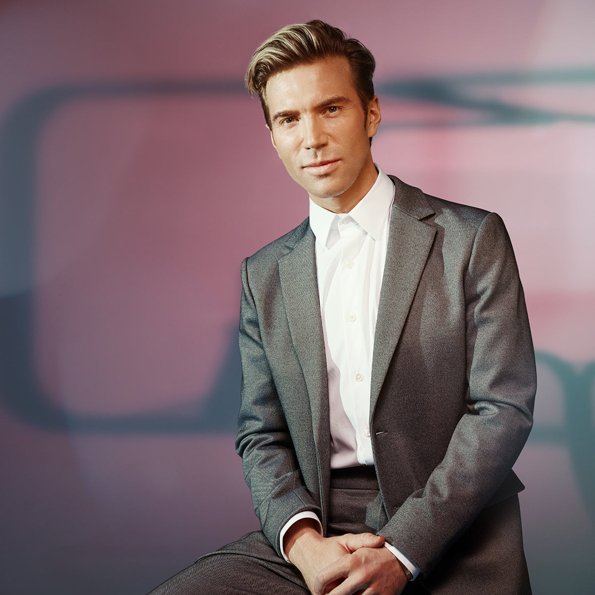 Portrait photo of Dr. Anthony, founder of Dr. Rossi DERM MD, seated on a stool and facing the camera, wearing a light grey suit with a white collared shirt, set against a pink and blue background. 